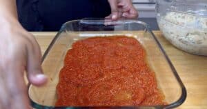 Glass baking dish with a layer of tomato sauce, ready for lasagna, next to a bowl of ricotta mixture on a wooden counter.